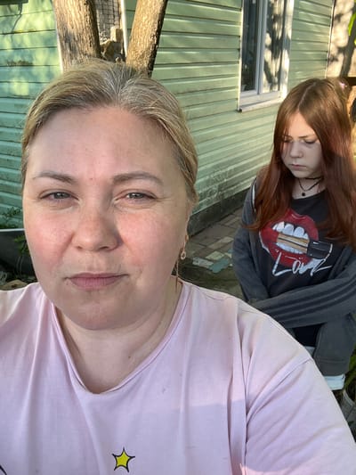 The image is a selfie of a fair-skinned woman with blonde hair. She is wearing a pink t-shirt with a yellow star. Behind her is a young woman with long auburn hair, a black choker necklace, and a black t-shirt with a red tongue and lipstick design under a gray hooded jacket. The background features a green building with a window and a tree. The location is Vesyoloye, Russia.