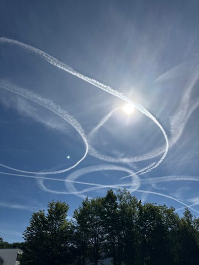 A bright sun shines in a clear blue sky above a line of dark green trees in Salach, Germany. Numerous white contrails from aircraft crisscross the sky, forming circular and linear patterns. The contrails are at various stages of dissipation, some appearing wispy and others more solid. The sun is partially obscured by a contrail, creating a lens flare effect. In the lower left corner, the top of a white building is visible.