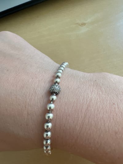 The image shows a close-up of a person's wrist with a silver beaded bracelet. The bracelet features several smooth, round silver beads, with one larger, textured bead in the center that appears to be encrusted with smaller stones or beads, giving it a sparkly appearance. The bracelet is resting on skin that shows fine lines and pores. The background is a light, neutral-colored surface, possibly wood or laminate, with subtle grain patterns visible. The lighting appears to be soft and diffused, casting no harsh shadows. The overall impression is one of a personal accessory being displayed or worn casually. There is no discernible text in the image. The scene is a simple, close-up shot focused on the jewelry and the wrist.
