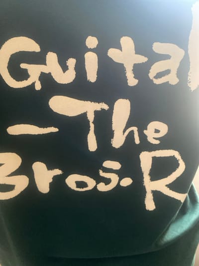 The image displays a close-up view of a dark teal or dark green textile surface featuring bold white lettering with irregular, brush-stroke-like edges. The text is arranged in three visible lines: the top line reads "Guital", the middle line shows a horizontal dash followed by "The", and the bottom line displays "Bros. R". A bright, diffuse light source is evident in the upper right portion of the frame, causing a glare across that area of the fabric. No individuals, additional objects, specific actions, or environmental context are discernible within the image. The location in the city of Katcha, Nigeria, cannot be determined from the visual information presented.