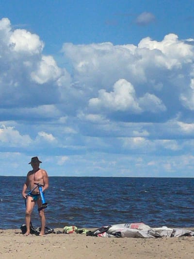 A person stands on a sandy beach in front of a body of water under a partly cloudy sky. The person is wearing a dark hat and blue swimming trunks, and is holding a blue and black hand pump. To the left of the person is a black bag, and to the right are several pieces of equipment, possibly related to water sports, including what appears to be a kite. The water is dark blue and rippling, and the sky is a bright blue with large, white, cumulus clouds. The scene is likely a daytime photograph. The location is the city of Belozersk, Russia, which is situated on the shore of Lake Beloye.