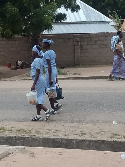 Two young women in matching light blue outfits and head coverings walk along a paved road in Biliri, Nigeria. Each carries a clear plastic bucket filled with food and a dark bag. In the background, a wall borders buildings with a metal roof. Another woman wearing a patterned head covering and long skirt walks in the opposite direction. A red container and a resting animal are visible near the wall.