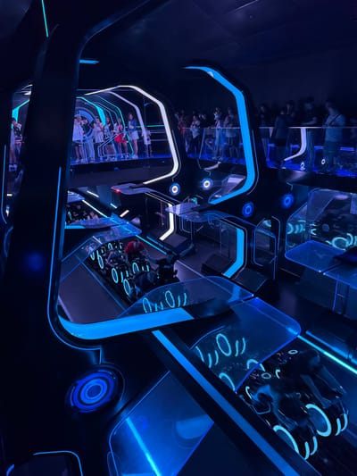 The image displays an interior view of the TRON Lightcycle / Run attraction, located at Magic Kingdom Park within Walt Disney World Resort in Orlando, Florida, United States. The scene is characterized by a futuristic aesthetic with predominantly dark surfaces illuminated by stark blue and white linear lighting elements.
On an elevated platform spanning the upper left and right portions of the frame, approximately 20-30 individuals are visible. These individuals, primarily adults, are standing behind transparent barriers or railings, observing the ride below. They are dressed in casual attire.
Below this observation level, several TRON Lightcycle ride vehicles are present on a track. At least two individuals are seated on these motorcycle-like vehicles, positioned in a forward-leaning riding posture; one rider is distinctly visible in a red shirt. The ride vehicles exhibit glowing blue circular accents. The track itself is dark with illuminated blue patterns. The surrounding architectural elements consist of dark panels with integrated blue and white light strips, creating a high-tech, digital grid appearance. The overall scene captures an active queue or viewing area of a theme park ride.
