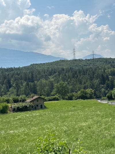 This is a vertical, wide-angle photograph taken outdoors on a sunny day, likely in the late morning or early afternoon. The scene depicts a rural landscape in Ossimo Inferiore, Italy.

The foreground is dominated by a lush green meadow dotted with small white flowers. To the left of the meadow, nestled at the edge of a dense, dark green forest, is a small, rustic stone house with a red-tiled roof and green-trimmed windows. A wooden staircase leads up to the house. Beside the house, a tree with green foliage is visible.

Behind the forest, a towering, mist-shrouded mountain range forms the backdrop. Power lines and two tall transmission towers are prominent against the distant, hazy blue mountains and the sky. The sky itself is a vibrant blue, filled with large, billowing white clouds, suggesting fair weather.

The overall impression is one of natural beauty and tranquility, with the human presence indicated only by the solitary house and the modern infrastructure of the power lines. There is no visible text in the image.