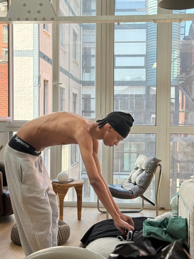 A young, lean male, shirtless and wearing light grey sweatpants with a visible black "Supreme" brand underwear waistband, is depicted indoors in Saint-Denis, France. He wears a black durag/beanie and has short dark braided hair. He is bent forward at the waist, with his arms extended downwards towards items on the light-colored floor. His hands are engaged with a black garment and other folded clothing, including a green item.

To the left, a dark brown leather armchair and a woven pouf are partially visible. A small wooden side table with a white spherical object on a coaster is positioned nearby. To the right, a grey cushioned lounge chair with a metal frame and red stitching details is present. A large clear glass bottle rests on the floor next to the chair.

The scene is brightly lit by natural light entering through large, multi-paned windows that form the background. The windows reveal an urban landscape consistent with Saint-Denis. Visible through the windows are brick buildings with white and black sections to the left, and modern glass-fronted structures to the right, including a distinctive cylindrical building with a ridged glass facade, which is identifiable as the Tour Pleyel. An industrial-style white metal beam with visible rivets spans the upper portion of the interior space.