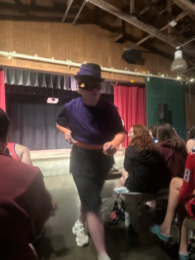 A person in a purple shirt, black shorts, a black hat, and goggles stands in a dimly lit indoor space with a wooden ceiling and walls. A stage with red curtains is visible in the background, suggesting a performance or event. Several other people are seated and standing around the main subject, some blurred due to motion. The overall scene appears to be casual, possibly a school or community gathering. The specific location in Washington Township, United States of America, cannot be determined from the image.