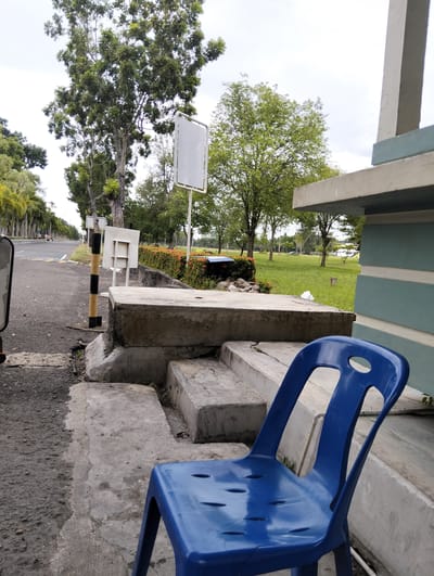 The image depicts an outdoor scene, likely in Tambontunong, Indonesia. In the foreground, a bright blue plastic chair sits on a concrete surface. Behind the chair, there are concrete steps and a low concrete wall. The background reveals a road with a black and white striped pole and a white sign. Beyond the road, a grassy area is dotted with trees. A building with a light blue and white facade is visible on the right side of the frame. The sky is overcast. There are no people visible in the scene.