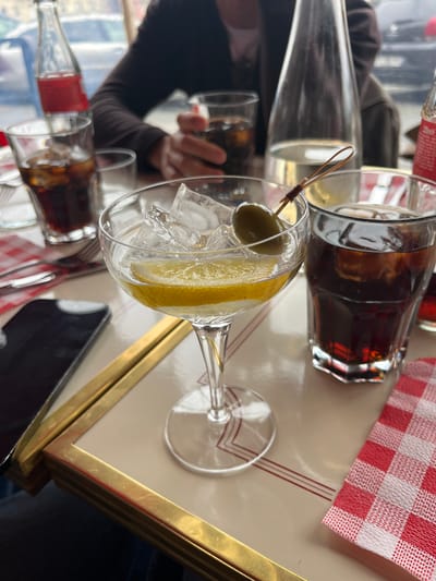 The image captures a scene at a table, possibly in a restaurant or bar, in Bayonne, France. A partially filled martini glass, containing ice, a lemon twist, and an olive on a toothpick, is in the foreground. Next to it are other glasses containing a dark liquid, presumably cola. There are bottles of cola as well. A red and white checkered napkin lies on the table. A smartphone sits to the right of the martini glass. A person with a dark-sleeved arm is seen reaching for one of the cola glasses in the background. The table is finished with a cream-colored top outlined with a gold trim. The environment is bright and suggests indoor lighting.