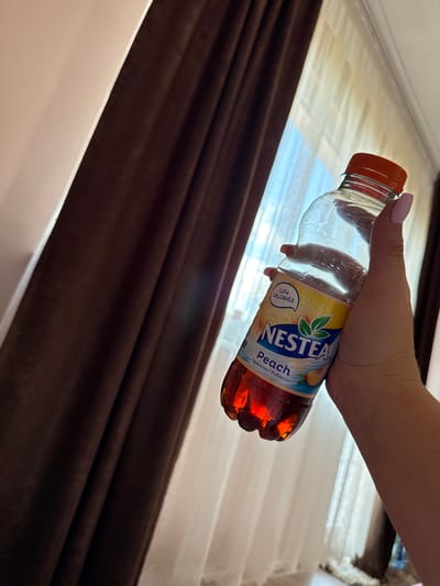 A person's hand, with light pink nail polish, holds a clear plastic bottle of Nestea Peach iced tea. The bottle is angled to the right, with the orange cap at the top. The label on the bottle features the Nestea logo in blue and white, with the word "Peach" in larger lettering, and a depiction of a peach. Below "Peach," there is text in a smaller font that appears to be in a Slavic language, possibly Bulgarian. The bottle is partially filled with a reddish-orange liquid. The background consists of dark brown curtains on the left and sheer white curtains with sunlight filtering through on the right. The scene is indoors, likely in a room with natural light. There are no discernable landmarks or elements that specifically place the image in Rogosh, Bulgaria.