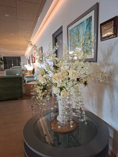 The image shows a still life arrangement within a modern interior setting, possibly a hotel lobby or lounge in Salou, Spain. The primary subject is a large, elaborate floral arrangement of white roses and delicate white and light purple filler flowers, artfully displayed in a vase on a round glass-topped table with a grey base. The table itself is placed on a wooden floor.

Above the table, a framed abstract artwork with predominantly blue and yellow tones hangs on a white wall. Another smaller, darker framed artwork is positioned higher on the wall, above the larger painting. In the background, the scene extends down a hallway lined with recessed lighting along the ceiling. A row of light blue upholstered seating and a dark green upholstered sofa are visible in the distance, suggesting a comfortable seating area. The overall ambiance is serene and sophisticated. There are no people visible in the image, and the time of day is indeterminate, though the artificial lighting suggests it could be evening or a dimly lit interior. There is no visible text in the image.