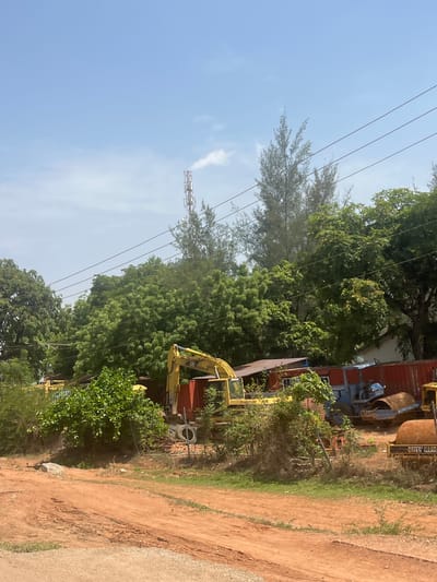 This outdoor image, captured in Galadimawa, Nigeria, presents a mix of natural landscape and an industrial or construction setting under a clear blue sky with minimal cloud cover, indicative of daytime.
The foreground and right side of the image feature a reddish-brown dirt road or path, with areas of green grass and a small grey rock visible on the ground. Abundant green foliage dominates the central and left portions, with various types of trees and bushes contributing to a lush environment. Some trees have needle-like leaves, while others show broader foliage.
Several power lines stretch across the upper left and central areas of the frame. In the distance on the left, a tall metallic lattice tower, likely a telecommunication or cell tower, rises against the sky, partly obscured by trees.
On the right side, heavy machinery is visible. A yellow excavator, bearing a partially visible "CAT" logo (indicating Caterpillar), is positioned behind some vegetation. Further back, a large red shipping container stands alongside other industrial equipment, including a prominent brown, drum-shaped object that appears to be a cement mixer. Stacked tires are also visible on the ground near these machines.
No individuals are present in the scene, suggesting a quiet moment or an inactive work area. The lighting indicates a bright, sunny day. Beyond the "CAT" logo, no other discernible text is clearly visible.