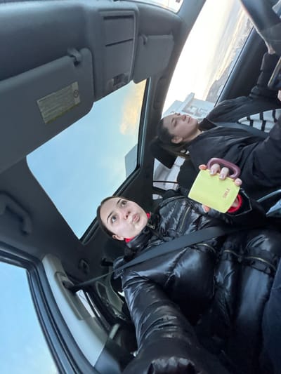 The image captures a casual scene inside a car, seemingly during the daytime in Hoenheim, France. The primary subject is a young woman, seated on the right side of the car, wearing a shiny black puffer jacket over a red garment, with a seatbelt visible across her shoulder. She holds a light yellow mug with a purple handle, on which the word "SMILE" is clearly legible. Her expression is neutral or slightly curious as she looks towards the camera.
In the upper-left portion of the image, the reflection of another young woman is visible, likely in an overhead car window or sunroof. She is wearing a black and white striped top under a black jacket and appears to be looking away. The car's interior features prominently, including parts of the headliner, sun visors (one displaying a warning label with small, indistinct text), and a side window.
Through the car's windows and reflections, a bright, clear to partly cloudy sky is visible, along with a distant building, indicating an outdoor, daytime setting. The overall mood is calm, depicting a paused moment, perhaps during a drive or while parked.