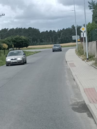 A silver Skoda Octavia estate car is parked on the left side of a paved road in Čistá, Czechia. The car is facing forward and its headlights are on. In the background, a dark grey sedan is driving away from the camera. A paved sidewalk runs along the right side of the road, bordered by a stone wall and vegetation. Traffic signs, including a diamond-shaped yield sign and a rectangular stop sign, are visible on the right side of the road. The sky is overcast, and the surrounding landscape consists of trees and fields.