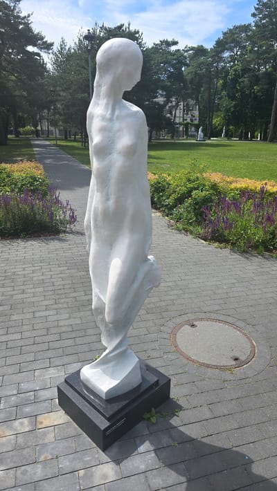 The image shows a white statue of a female figure in a park setting. The statue is located on a paved walkway with grey bricks. The figure is abstract, with smooth, flowing lines, and is posed with one leg bent. It stands on a dark rectangular base with an inscription. Behind the statue, there is a well-maintained lawn with scattered trees and flower beds containing purple and yellow flowers. In the background, there are more trees, a building, and other statues. A manhole cover is visible on the walkway to the right of the statue. The sky is blue with scattered white clouds. The scene appears to be in Konstancin-Jeziorna, Poland, likely in a public park or garden, given the presence of sculptures and manicured landscaping.