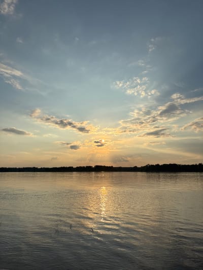 The image depicts a serene sunset over a wide body of water, likely a river, with a treeline on the distant horizon. The sun is partially obscured by clouds, casting a warm, golden light that reflects on the rippling surface of the water. The sky is a gradient of blue and orange, with scattered clouds. The water in the foreground is calm, with subtle waves. The scene is devoid of any human presence or man-made structures, suggesting a natural, undeveloped landscape. The location is identified as the city of Ol'khovskoye, Russia.