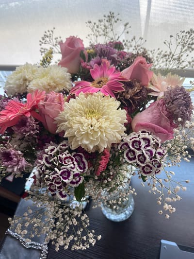 The image shows a close-up of a flower bouquet, featuring a variety of blooms including pink roses, white chrysanthemums, pink gerbera daisies, purple carnations with white edges, and baby's breath. The arrangement is dense and colorful. A vase containing the flowers is partially visible, and a window with sheer curtains is present in the background. The surface on which the bouquet sits is dark and appears to be wood. The image does not provide sufficient information to confirm the location in the city of Barrio de Nuevo León, Mexico, and does not depict any people, actions, or interactions.