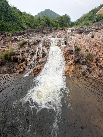 A wide-angle, eye-level shot captures a waterfall cascading over rocky terrain at a location in the city of Khanh Vinh, Vietnam. The waterfall is the primary subject, with white, foamy water tumbling down a series of layered, reddish-brown rocks. The rocks are uneven and textured, with some areas showing moss or small green plants growing from crevices.
The water at the base of the waterfall is dark and appears turbulent, with white foam swirling on the surface. The surrounding environment is lush and green, with dense vegetation covering the hillsides that rise steeply on either side of the waterfall. The trees are a deep green, and the foliage is thick, suggesting a tropical or subtropical climate.
In the background, the sky is overcast, with a soft, diffused light. In the upper right corner of the image, a portion of a simple, open-sided shelter or canopy structure is visible, supported by metal poles and with a light-colored roof. This structure suggests that the area is a tourist or recreational spot.
The overall scene is one of natural beauty, with the powerful force of the waterfall contrasting with the serene green landscape. The image appears to be taken on a day with no direct sunlight.