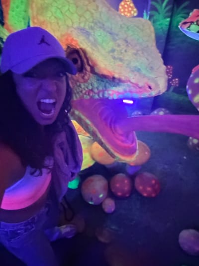 In a dark, possibly indoor, environment illuminated with blacklight, a person wearing a dark blue baseball cap, a shirt, and what appears to be jeans is lying down and looking up with their mouth open in a possible smile or shout. Above them is a large, brightly colored sculpture of a dinosaur head and a large mushroom. There are other similar mushrooms in the background. The location is uncertain and there's no clear evidence to confirm the location of Binh Lu, Vietnam.