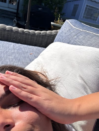 An individual is depicted reclining outdoors on a patio, shielding their face from direct sunlight with their right hand, eyes appearing closed or partially closed. They are resting on a modern outdoor sofa featuring dark grey woven wicker and denim-blue cushions, with a light grey pillow supporting their head. In the background, to the left, a black and white dog is visible lying on a stone or paved surface near a building with a glass door. A black Weber grill, a patio umbrella pole, and potted plants are situated in the mid-background. Further right, a blue canvas tent-like structure with white trim is partially visible. The scene is brightly illuminated by strong sunlight, indicating a clear day. This scene is situated in the city of Princeton, United States.