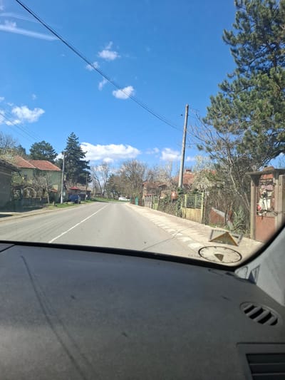 View from inside a vehicle, looking out onto a paved road in what appears to be a residential area. The road has a white line marking the center. Houses with red tiled roofs are visible on both sides of the road, partially obscured by trees and greenery. Utility poles and power lines run alongside the road. In the foreground, part of the vehicle's dashboard is visible, including vents and a small clock or gauge mounted on the dashboard. The sky is blue with scattered clouds. Based on visual characteristics, this is Studeno Buche, Bulgaria.