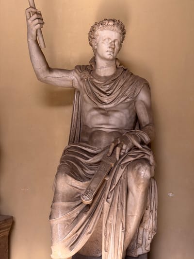 A full-length marble statue of a seated male figure is depicted against a plain, textured light brown wall. The figure wears a laurel wreath on his head and a draped garment that covers his lower body and left shoulder, leaving his right shoulder and chest bare. His right arm is raised, holding a plain, cylindrical staff. His left hand rests on his lap, grasping a wide, flat object with decorative carvings, possibly a sword sheath or a scroll holder. The white marble exhibits numerous cracks, particularly visible on the arms, torso, and legs, along with areas of reddish-brown discoloration. The lighting is soft and even, originating from the upper left. This scene is located in Città del Vaticano, Vatican City.