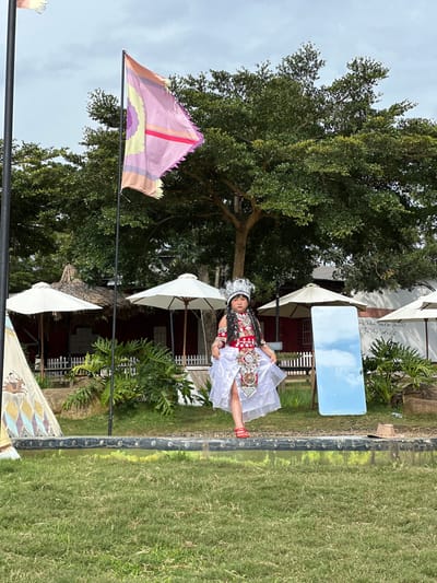 A young girl, dressed in a traditional white and red outfit with an elaborate headdress, stands in front of a red building with thatched-roof awnings and white umbrellas. She is positioned on a raised platform overlooking a grassy area and a reflective pool. Behind her, a large tree provides shade, and a flag with pink and yellow stripes flutters in the wind. To her right, a tall mirror reflects the sky. A cowboy hat lies on the ground near the edge of the platform. The setting appears to be a recreational or cultural space within Thôn 5, Vietnam, characterized by lush greenery and rustic structures.