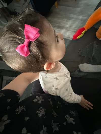 The image presents a close-up, tilted view of a young child and parts of an adult within an indoor setting. The central subject is a child, likely a toddler, seen from a high angle focusing on the top of their head and the right side of their face. The child has light brown hair adorned with a vibrant pink, ribbed bow hair clip positioned on the left side of their head. They are wearing a light-colored, possibly cream or white, long-sleeved top featuring small, repetitive dark patterns, which resemble scissors or tiny bows. The child's right ear, cheek, and a portion of their right eye, looking upwards, are visible.
An adult is partially visible to the right and behind the child, wearing dark, possibly black, clothing with grey or white abstract floral patterns. The adult's arm is seen extending towards the child, suggesting they are holding or supporting the child.
In the blurred background, to the upper left, an orange and red plush toy, possibly a character or a carrot, is discernible. Further back, grey fabric, likely part of clothing or a blanket, is visible alongside darker furnishings. A light-colored, possibly wooden, surface appears in the background.
The scene depicts a calm, intimate moment, suggesting an interaction or cuddle between the child and adult in a domestic environment. The lighting is ambient, indicating an indoor setting but not providing specific cues about the time of day or weather. There is no visible text in the image. The location context provided is Macclesfield, United Kingdom.