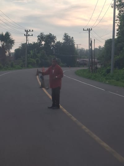 A man in a red shirt and dark pants stands in the middle of a two-lane asphalt road, possibly near Keude Lueng Putu, Indonesia, as indicated by the surrounding vegetation and utility poles. The man is holding a bottle in his right hand and appears to be placing it down on an orange traffic cone. Another traffic cone is visible nearby. The road has a yellow line down the center and a white line on the right edge. Utility poles with electrical wires are present on both sides of the road, and trees and lush green foliage line the sides of the road. The sky is overcast with a pale, diffused light, suggesting it is either early morning or late evening. The overall scene depicts a potentially hazardous situation, with a person obstructing traffic on a roadway.