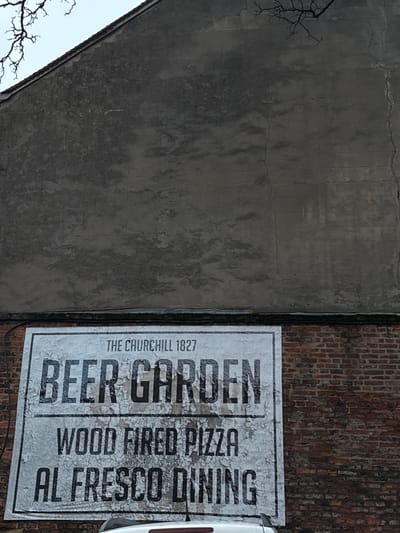 A large rectangular sign on a brick wall advertises "Beer Garden, Wood Fired Pizza, Al Fresco Dining." The sign has a distressed, vintage look with peeling paint. The text is bold and blocky. The words "The Churchill 1827" are printed above "Beer Garden." To the left, a large concrete wall with cracks and water stains dominates the frame. A bare tree branch extends into the lower left corner. A vehicle is partially visible on the right.