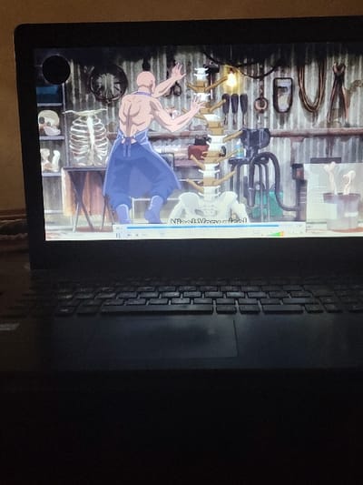 The image displays a black laptop screen, located in the city of Kubare, Nigeria, showing an animated scene. The animation depicts a bald, muscular male figure, viewed from the back, wearing a blue apron and light blue lower garments, with arms raised and hands open towards a large, stylized white spinal column model with yellow/brown sections and a pelvis at its base. The animated background is a workshop or laboratory with a corrugated metal wall. To the left, a human rib cage model and skull rest on a table, with shelves containing additional objects. To the right, various tools, including hooks and chains, hang on the wall, illuminated by a glowing light bulb. A video player interface, including a progress bar and partially visible text ("Nioa!! Mary nice!"), overlays the bottom of the animated scene. The physical laptop's dark keyboard and touchpad are visible in the foreground, under low ambient light.