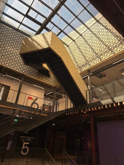 The image captures an interior view of a modern, multi-level building with a striking architectural design. A golden, perforated staircase is suspended from the ceiling, dominating the central space. The ceiling itself is a vast expanse of glass panels supported by dark metal beams, allowing natural light to filter in. Below the suspended staircase, another set of open-tread staircases with metal mesh railings are visible, leading to different levels. The walls are a mix of white plaster and dark metallic accents, with large, stylized numbers "6" and "5" painted on them, and signage indicating "Tijdelijke tentoonstelling Temporary exhibition." This interior appears to be part of a cultural institution or museum. The architectural style suggests a contemporary building, possibly located in a city known for modern design. Based on the context of the numbers and exhibition signage, it is likely an indoor exhibition space.
