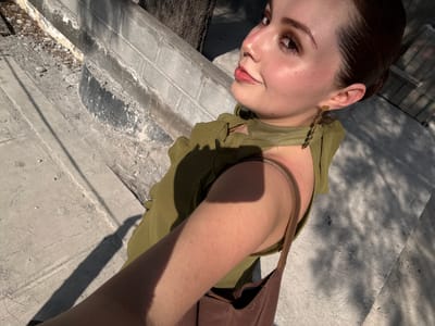 A young woman is shown from a low angle, looking upward. She has fair skin, brown hair pulled back, and is wearing a green top with a brown strap from a bag or garment over her shoulder. She is wearing an earring on her left ear. The background consists of concrete and brick surfaces, with shadows. The image was taken in San Pedro Garza García, Mexico.