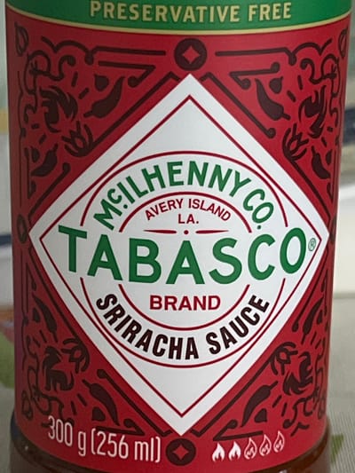 A close-up, rotated shot captures the label of a Tabasco Brand Sriracha Sauce bottle. The label is predominantly red with intricate dark red patterns featuring chili peppers and abstract swirls. A central white diamond-shaped area prominently displays the product information.
Visible text on the label includes "PRESERVATIVE FREE" in green along the left edge. The main brand identity, "MCILHENNY CO. AVERY ISLAND LA. TABASCO BRAND SRIRACHA SAUCE," is displayed in a circular green font around the "TABASCO" logo, which is vertically oriented. Below this, "SRIRACHA SAUCE" is horizontally aligned. Further to the right, the weight and volume are indicated as "300 g (256 ml)." The background is blurred, suggesting a shallow depth of field, bringing the product label into sharp focus. The overall impression is a vibrant and detailed product advertisement.