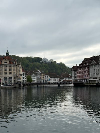The image depicts a waterfront view in Lucerne, Switzerland. The Reuss River flows between historic buildings. On the right, buildings with timber framing and white facades are visible. Across the river, more structures are built into a hillside covered with dense green trees. A bridge with dark metal railings spans the river. A white building with a dome, likely the Chateau Gütsch hotel, sits atop the hill in the distance. The sky is overcast and gray, reflecting in the water. The water shows ripples and reflections of the buildings. At the bottom of the frame, a building with a green-domed tower is partly visible.