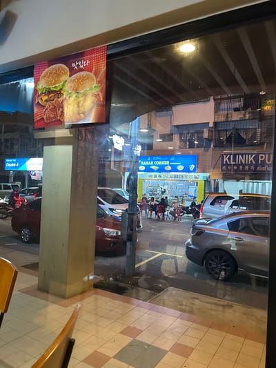 The image presents a nighttime view from inside a building, looking out through a glass window onto a commercial street within the Subang Jaya City Council, Malaysia. Reflections are evident on the glass, including parts of a wooden chair in the bottom left, a tiled floor with light and pinkish-brown squares, and a large red poster displaying two hamburgers and fried chicken, accompanied by Korean text "맛있다" (delicious) in the upper-left. A thick, light-colored square pillar is prominent on the left side, extending from the bottom to the mid-frame.
Beyond the glass, the street scene reveals various businesses and activities. On the far left, a blue sign for "KEDAI ALAT TULIS Double A DDX SOLUTIONS" is visible above a parked red sedan, near which a person on a motorbike is situated. Further right, a brightly lit food stall named "RAZAK CORNER ENTERPRISE" (indicated by a blue sign with Arabic script and English text) features several individuals dining at red and black tables placed on the sidewalk. Motorcycles are parked near the stall. To the right of "RAZAK CORNER," a blue sign for "KLINIK PU" displays multilingual text in English, Chinese (内外骨科跌打 跌打), and Tamil (உள் வெளி எலும்பு மூட்டு சிகிச்சை), indicating an orthopedic clinic.
Several vehicles are present on the street; a silver car with a Malaysian flag sticker on its rear window is partially visible behind a dark grey sedan with black rims in the bottom right foreground. The buildings across the street are multi-storied, exhibiting a mix of older and newer architecture with visible windows and balconies. The entire scene is illuminated by a combination of streetlights and lights from the businesses, characteristic of an active urban environment after dark.