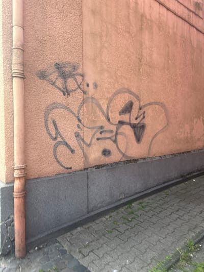 The image depicts an outdoor scene in the city of Gelsenkirchen, Germany. A roughly textured, peach-colored wall dominates the frame, showing signs of wear and discoloration with some greenish-grey streaks. To the left, a vertical, rusty orange drainpipe runs along the wall. The lower portion of the wall is covered by a dark grey, textured panel. Below this, a section of interlocking grey pavers forms a walkway, with sparse green weeds growing in the cracks.
Graffiti in dark grey spray paint is visible on the peach-colored wall. On the left side of the wall, there is abstract, sprawling graffiti that appears to form a stylized, angular shape with some interconnected lines. To the right of this, another piece of graffiti consists of large, rounded, looping shapes. Further to the right, a distinct graphic element resembling a stylized bird or face with a triangular shape in its mouth is present. There are also smaller, less defined spray-painted marks and smudges on the wall. The lighting suggests it is daytime, with a soft, diffused light.