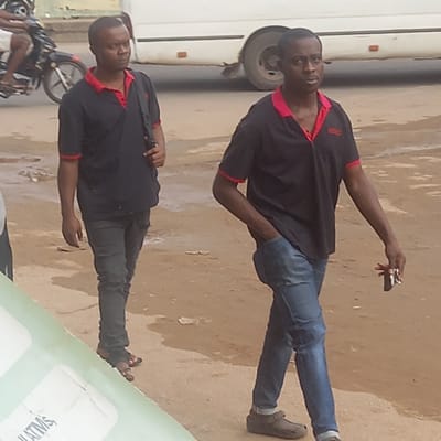 The image depicts a man in the city of Sardauna, Nigeria. He is wearing a black polo shirt with red trim, blue jeans, and brown sandals. He is standing near a white vehicle and a parked motorcycle. He is holding a small object in his raised hand. The background shows a sandy or dirt surface, typical of a street in Sardauna. A partial view of what seems to be a sign is on the right side.