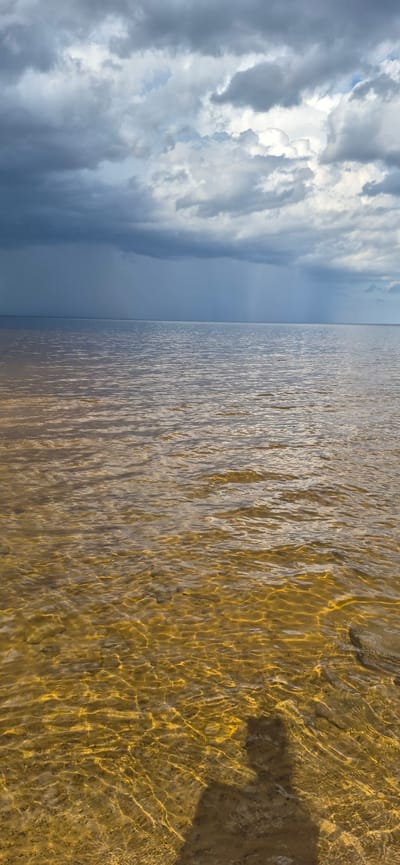 The image shows a body of water with a sandy bottom, viewed from a low angle at the edge of the water. The water is shallow and clear, with sunlight filtering through to illuminate the sand and ripples. The sky above is dramatic, filled with dark, heavy clouds that suggest an approaching storm. A faint rain can be seen falling in the distance. The shadow of a person, presumably the photographer, is cast on the sandy bottom in the foreground. There are no other people or objects visible in the scene, and no specific landmarks are identifiable that would place the image in Motornoe, Russia.