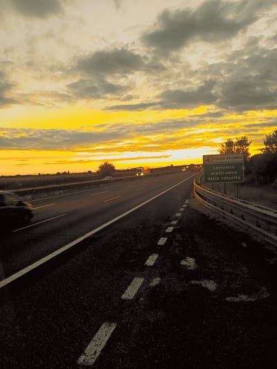 A bright yellow and orange sunset casts an intense glow across a cloudy sky over a highway in the city of Somaglia, Italy. The highway is dark asphalt with white lane markings and a metal guardrail. On the right side of the highway, a green sign with white text reads "Polizia Stradale controllo elettronico della velocità," indicating a police speed check. There are vehicles on the highway, appearing as blurred shapes due to motion. The surrounding landscape is dark with silhouetted trees and fields. The overall scene depicts a sunset drive on an Italian highway.