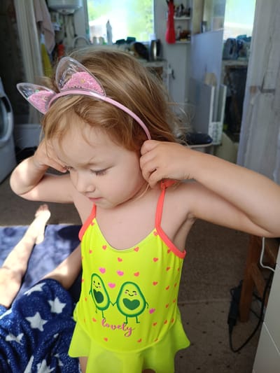 A young girl with blonde hair, wearing a neon yellow swimsuit adorned with pink hearts and two smiling avocado characters, stands indoors. She is adjusting a pink headband with clear, glitter-filled cat ears on her head. Her expression is thoughtful and her gaze is directed downwards.

In the background, a window shows a bright, daylight scene with greenery outside, suggesting it is daytime. The interior setting appears to be a room with various objects, including what looks like a washing machine on the left and shelves with items in the distance. A blue blanket with white stars is visible on the floor to the girl's left, with a person's legs and feet resting on it.

The text "lovely" is visible on the girl's swimsuit. The overall impression is one of a casual, perhaps playful, moment captured indoors.