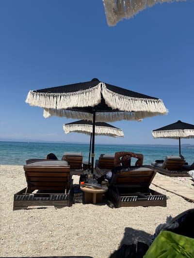 The image shows a beach scene on a clear, sunny day, likely in the afternoon given the bright sunlight and shadows. The foreground is dominated by two large beach umbrellas with black and white striped canopies and long, white fringe. The umbrellas are set up over wooden beach chairs.
In the middle ground, several people are visible. One person is lying on a beach chair under an umbrella, their arms resting behind their head, appearing relaxed. Another person's head is visible peeking over the back of a beach chair further down. The beach is covered in small pebbles and sand. A wooden boardwalk is partially visible.
The background features the calm, blue sea extending to the horizon, where a hazy coastline or mountains are visible. The sky is a clear, vibrant blue. There is no visible text in the image. The overall atmosphere is one of leisure and relaxation on a warm summer day at the beach.