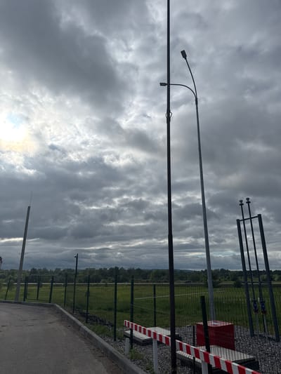 The image captures an outdoor scene under a dramatic, heavily clouded sky, with shades of grey dominating the upper and central portions. Patches of brighter light, suggesting the sun, are visible breaking through the clouds towards the lower left. The foreground features a paved path or road with a concrete curb running horizontally across the bottom.
Several tall, slender utility poles or light posts are prominent subjects, extending vertically across the frame and appearing silhouetted against the sky. One significant pole in the mid-left has two curved lampposts attached. Further to the right, a security fence, likely made of metal mesh, separates the paved area from an expansive green field. A red and white striped barrier post is visible on the right, positioned next to a red rectangular utility box. Additional metal structures, including one with horizontal bars and insulators, are seen near the fence line on the right. Small, dark security cameras (CCTVs) are mounted on some of the poles and fence posts, indicating surveillance.
In the background, beyond the green field, a dense line of trees or forest stretches along the horizon, with hints of distant, undulating terrain. No people or specific activities are depicted, giving the scene a quiet, almost contemplative atmosphere. The heavy cloud cover suggests an overcast day, possibly late afternoon or approaching evening. There is no discernible text visible in the image. The overall impression is that of a secured or infrastructure-heavy area set within a natural, rural or semi-rural landscape in Гремяча, Russia.