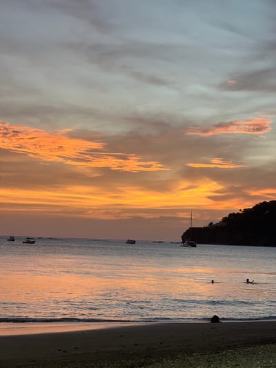 A serene sunset casts a warm orange and pink glow across the sky and the calm waters of San Juan del Sur, Nicaragua. The beach in the foreground is dark sand with small pebbles. Several boats are visible in the distance, some anchored and others moving. A few people are in the water, one appearing to swim. A dark, tree-covered cliff face rises on the right side of the image, with a few buildings visible at its summit. The overall scene is peaceful and picturesque, typical of a tropical evening.