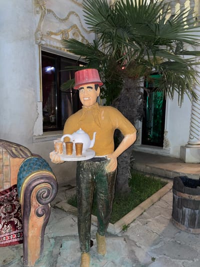 This is a full-body photograph of a life-sized statue of a man, likely a decorative garden gnome, wearing a red hat, a yellow shirt, and green pants. The statue is positioned outdoors in what appears to be the courtyard of a building. The man is holding a tray with a white teapot and four small glasses. To the left of the statue is a portion of another decorative structure, possibly a bench, with intricate carvings and a red patterned rug. In the background, there is a palm tree and a building with decorative stucco work. The ground is paved with stones, and there is a small patch of grass. A wooden barrel is visible to the right of the statue. The scene is lit, suggesting it is nighttime or dusk. The statue is located in the city of Kaspiysk, Russia.