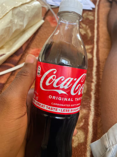 This media file shows a close-up of a person's hand holding a glass bottle of Coca-Cola. The bottle is mostly filled with dark liquid, and the iconic red Coca-Cola label is clearly visible. Text on the label includes "Coca-Cola," "ORIGINAL TASTE," "CARBONATED SOFT DRINK," and "LESS SUGAR." Nutritional information is also visible, stating "per 5 cl," "Energy 75 kca," and "50 cl." The background suggests an indoor setting, possibly a home, with a brown patterned blanket and a light-colored plastic bag. The lighting suggests it's daytime. The scene is casual, with the person's hand gently gripping the bottle. The focus is on the product and the hand holding it. The presence of "LESS SUGAR" indicates this is a variant of the classic Coca-Cola. The context provided mentions Wai A, Nigeria.