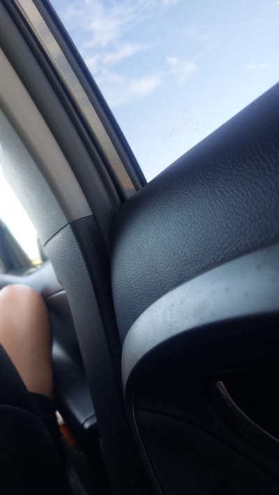 The image is taken from inside a vehicle, looking upwards and outwards through an open sunroof. The sky is visible, with some white clouds scattered across a bright blue expanse, suggesting it is daytime, likely sunny. The interior of the car is predominantly black, with textured surfaces on the roof lining and dashboard.
The primary focus is the contrast between the dark interior and the bright sky. A textured, dark material, likely leather or a synthetic equivalent, forms the edge of the sunroof opening and curves around the interior of the car. There is a metallic trim piece visible along the edge of the sunroof opening. In the upper right quadrant of the image, a dark, curved object, possibly part of the dashboard or steering wheel, is visible in shadow.
Further down and to the right, a person's leg and foot are visible, suggesting someone is seated in the car. The leg is clad in dark clothing. The presence of the sunroof implies the car is in a location where it can be opened, and the view of the sky suggests an outdoor setting. No specific time of day is definitively indicated beyond "daytime" due to the bright sky. No emotional cues are discernible from the image.
There is some faint, white text visible on the glass of the sunroof, near the top edge of the image. It appears to be a circular emblem with some lettering inside and "CB211B" written below it. This is likely a manufacturer's mark or safety certification for the glass. The overall impression is a relaxed, everyday moment inside a car on a clear day.