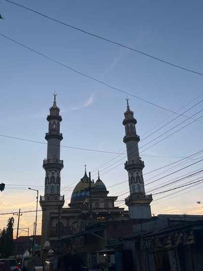 The image depicts a mosque in Balonggabus, Indonesia, with its prominent minarets and domes silhouetted against a dusky sky. Electrical wires crisscross the frame, obscuring parts of the sky and the mosque's architecture. In the foreground, a street scene unfolds with buildings and shops, their facades displaying various signs. A street lamp is visible to the left, and trees line the background. The overall impression is one of a bustling urban environment at dusk, with the mosque serving as a central architectural feature.