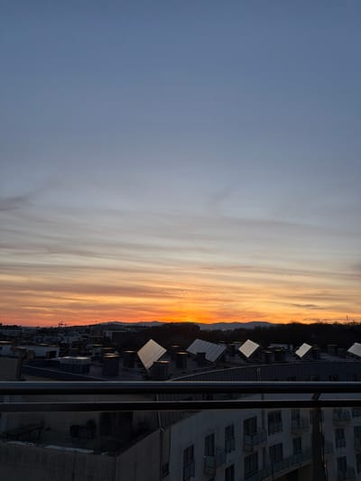 A cityscape stretches along the right side, featuring a multi-story building and numerous smaller structures. Air conditioning units are visible on the rooftops. The sky transitions from a vibrant orange at the horizon to a pale blue above, adorned with wispy clouds. The image is presented in a vertical orientation.