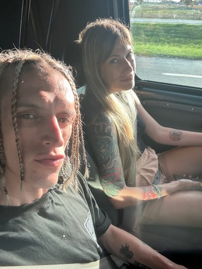 A low-angle shot shows two people lying down in what appears to be a vehicle.
The person closest to the camera is male, with fair skin and a hairstyle consisting of small braids. He is wearing a dark t-shirt and is lying on his back.
Behind him is a woman with blonde hair and visible tattoos on her arms and body. She also appears to be lying down. Outside the window, there is vegetation, suggesting a roadside environment. The location cannot be determined as Adela, Argentina.