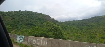 The image shows a view of a lush, green, hilly landscape under a cloudy sky. The foreground features a concrete barrier with graffiti. The graffiti includes text such as "HASKE ZIPHION", "4 BEARS", and "PKK". The hills are covered in dense vegetation, with some rocky outcrops visible. The overall scene suggests a natural, somewhat rugged environment, possibly in a rural or semi-rural area. Based on the graffiti's mention of "PKK", which is a militant group often associated with Nigeria, and the general landscape, this scene is likely located in Gu, Nigeria.